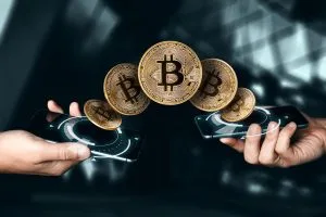 Zwei Hände halten Smartphones mit digitalen Schnittstellen, und fünf schwebende Bitcoin-Symbole scheinen zwischen den Geräten hin und her zu wandern, was für den Austausch von Kryptowährungen steht. Der moderne, verschwommene Hintergrund deutet die Frage an: Was ist Bitcoin?.