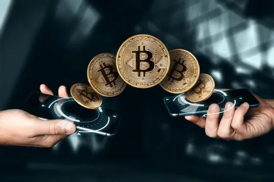 Zwei Hände halten Smartphones mit digitalen Schnittstellen, und fünf schwebende Bitcoin-Symbole scheinen zwischen den Geräten hin und her zu wandern, was für den Austausch von Kryptowährungen steht. Der moderne, verschwommene Hintergrund deutet die Frage an: Was ist Bitcoin?.