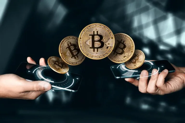 Zwei Hände halten Smartphones mit digitalen Schnittstellen, und fünf schwebende Bitcoin-Symbole scheinen zwischen den Geräten hin und her zu wandern, was für den Austausch von Kryptowährungen steht. Der moderne, verschwommene Hintergrund deutet die Frage an: Was ist Bitcoin?.