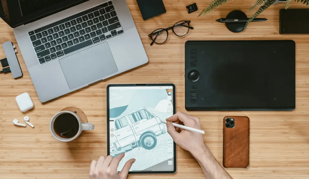 Draufsicht auf die Hände einer Person, die mit einem Stift NFTs auf einem Tablet zeichnet, umgeben von einem Laptop, einem Smartphone, Kaffee und Büromaterial auf einem Holzschreibtisch.