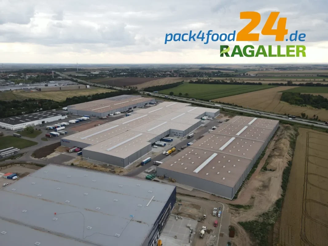 Luftaufnahme eines Industriekomplexes mit großen Lagerhallen, gekennzeichnet mit dem Logo und dem Text „pack4food24.de RAGALLER“ im Himmelsabschnitt. Umliegende Felder und ein Blick auf Magdeburg sind sichtbar und heben das neue Logistikzentrum hervor.