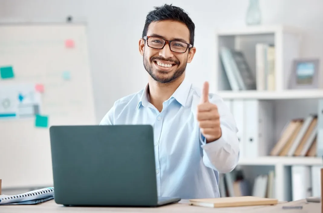 Ein Mann mit Brille sitzt an einem Schreibtisch mit Laptop, lächelt und zeigt mit dem Daumen nach oben. Im Hintergrund sind Regale mit Büchern und ein Whiteboard zu sehen, was an ein Büro bei Limtronik erinnert.