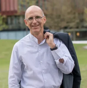 Warum setzt ein Rhetoriktrainer mit 35 Jahren Erfahrung plötzlich auf KI? Peter Flume mit Brille und hellem Button-up-Hemd steht im Freien, lächelt in die Kamera und hält eine dunkle Jacke über die Schulter. Der erfahrene Rhetoriktrainer ist vor einem unscharfen Gebäude und Gras im Hintergrund zu sehen.