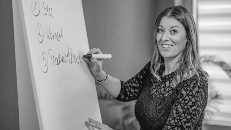 Sandra Agerer steht neben einem Flipchart, hält einen Marker und lächelt in die Kamera. Auf dem Flipchart ist eine nummerierte Liste zu sehen, was darauf hindeutet, dass sie in einem Büro oder Besprechungsraum einen Vortrag über Digitale Markenstrategie hält.