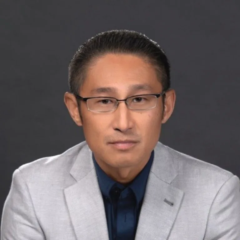 Joseph Kim mit kurzen dunklen Haaren, der eine Brille, einen hellgrauen Blazer und ein dunkles marineblaues Hemd trägt, sitzt vor einem schlichten dunklen Hintergrund und blickt mit neutraler Miene in die Kamera - er repräsentiert die innovative Agentic AI-Plattform von DRUID AI.