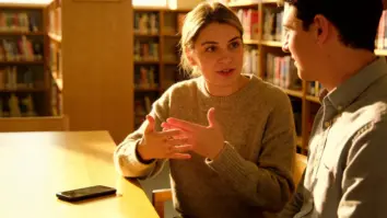 Eine gehörlose Frau gebärdet in einer hellen Bibliothek mit einem Freund, während ein unbeschriftetes Smartphone bereitliegt.