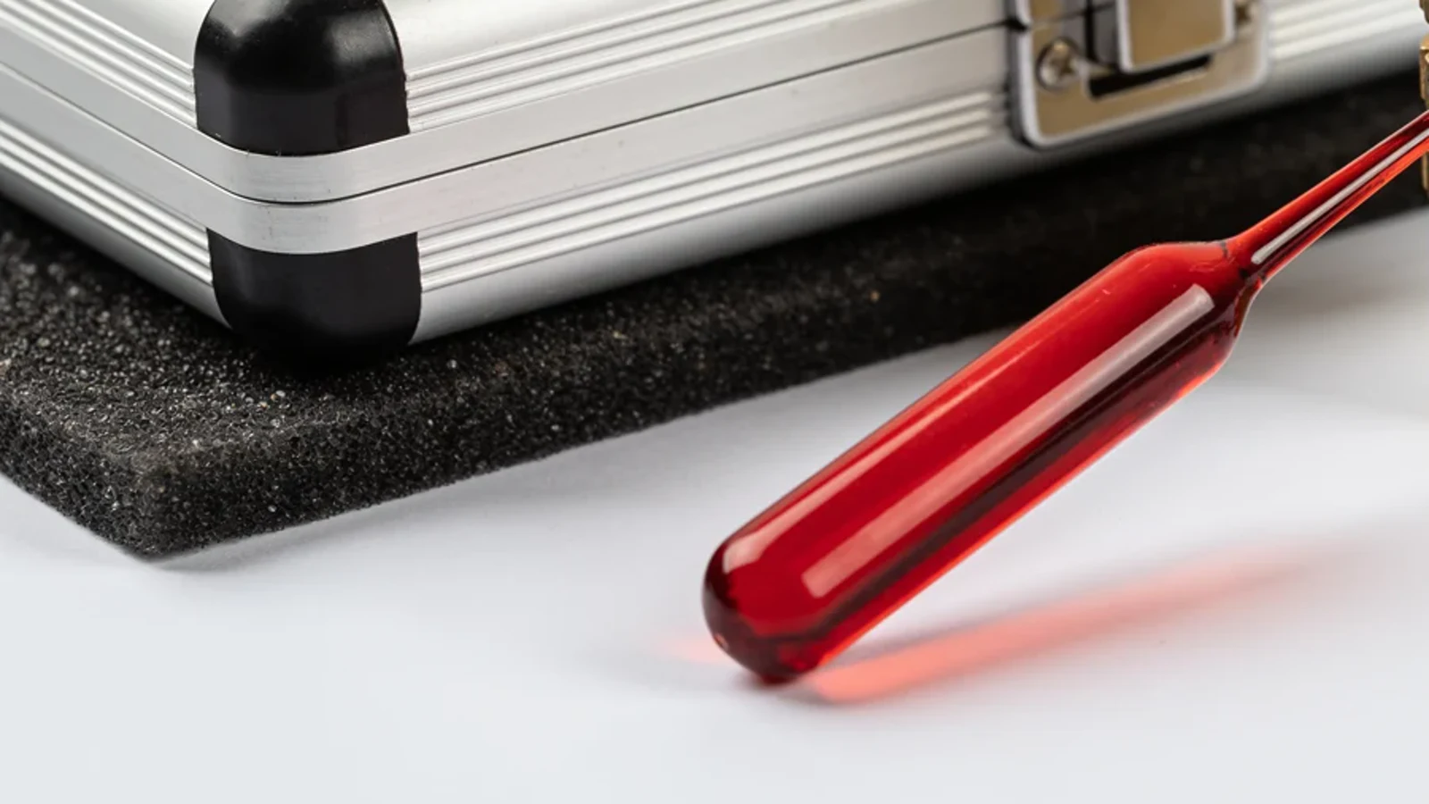 Eine rote Glasspitze durchbohrt einen Messing-Schlüsselmechanismus, während Metallglieder zu einer Schutzkassette hin abbrechen.