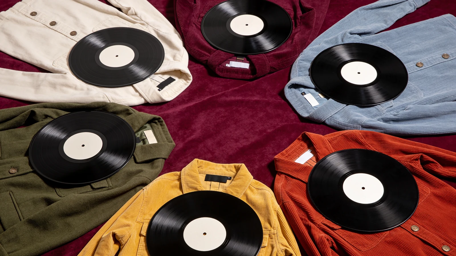 Sechs unterschiedliche Vintage-Kleidungsstücke liegen jeweils neben einer unbeschrifteten Schallplatte auf pflaumenfarbenem Stoff.