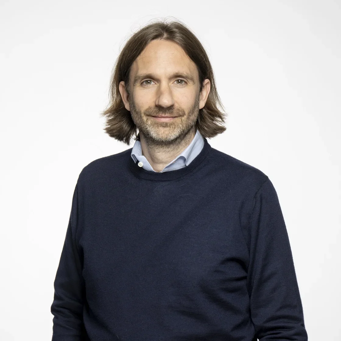 Michael Plankensteiner mit schulterlangen braunen Haaren und Bart steht vor einem schlichten weißen Hintergrund. Er trägt einen marineblauen Pullover über einem hellblauen Kragenhemd und verkörpert damit den professionellen Stil von Neuron Automation.